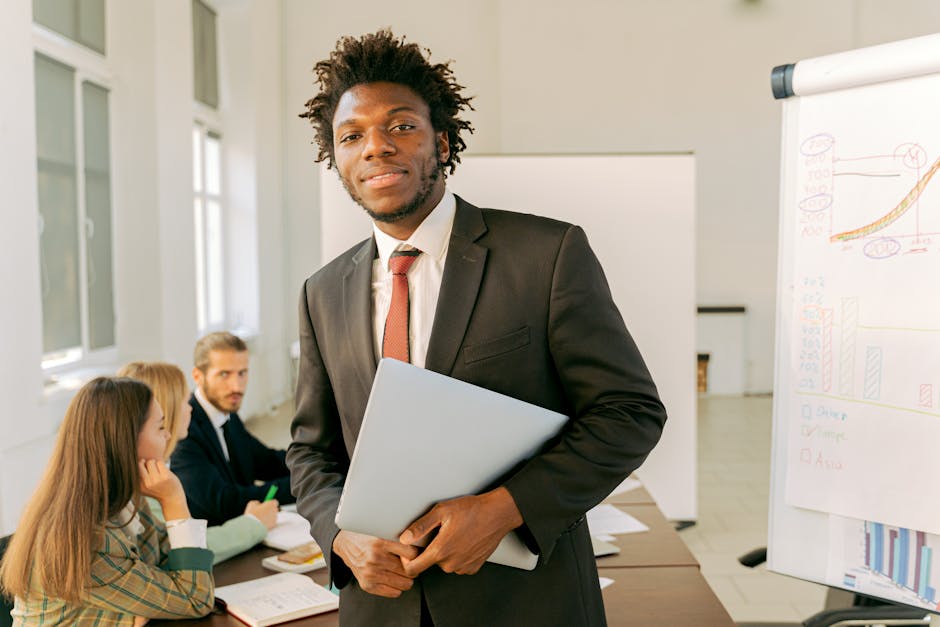 A confident businessman in a black suit holds a laptop during a meeting in an office setting